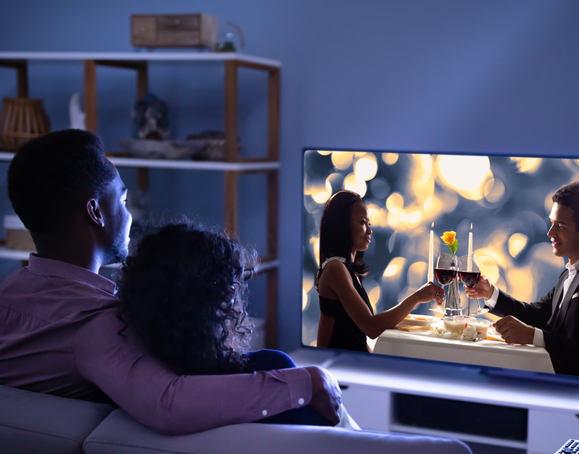 Imagem de um casal sentados em um sofá de frente para Smart TV 50” DLED 4K Multi assistindo 2 pessoas brindarem uma taça de vinho