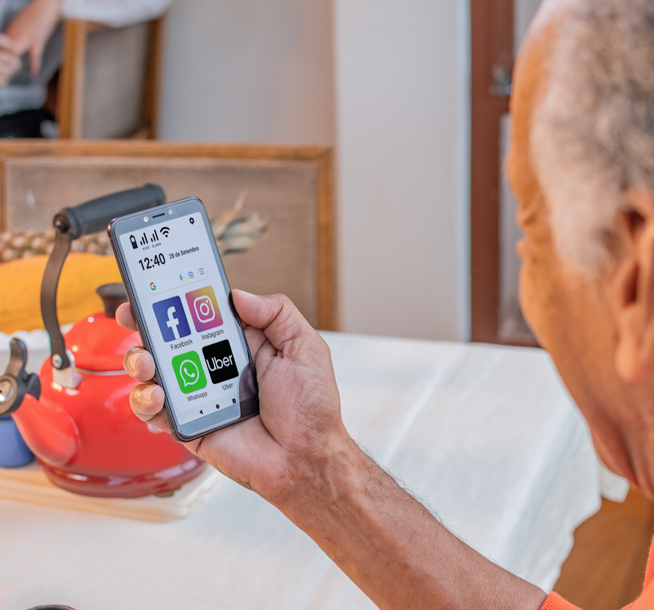 A imagem mostra um senhor olhando para um smartphone que tem na tela ícones grandes de aplicativos populares como Facebook, Instagram e Uber. O design sugere facilidade de uso para usuários mais velhos.