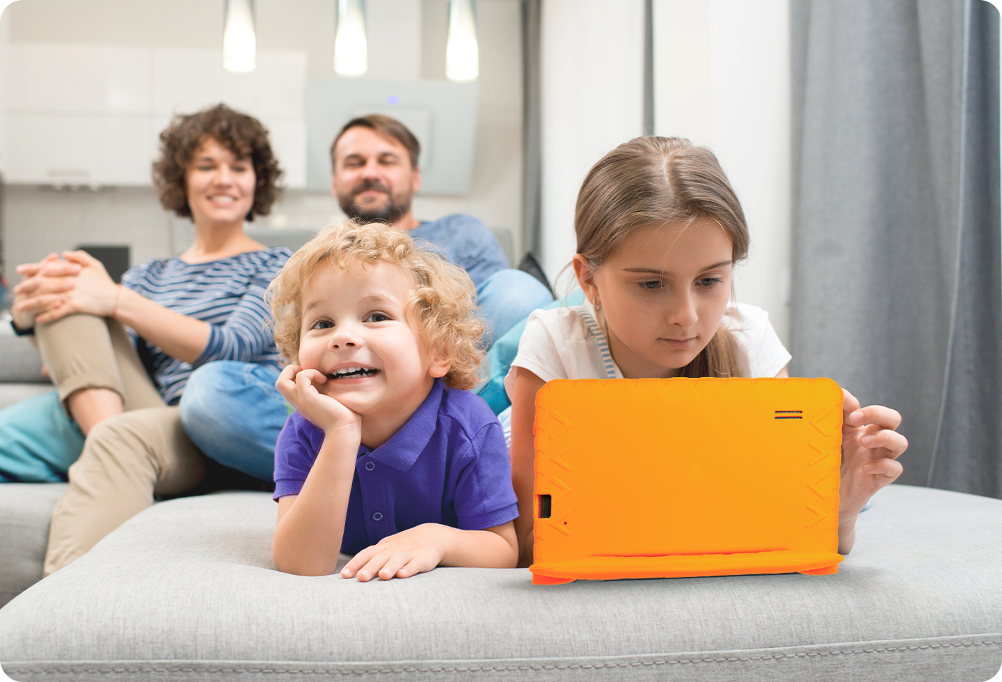 Foto de uma família alegre sentada em um sofá numa sala de estar, com um menino e uma menina deitados de barriga para baixo, sendo que a menina está usando o Tablet Kid Pad.