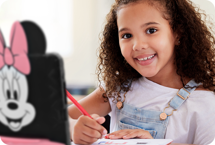 Criança sorridente e desenhando algo, enquanto há um Tablet Minnie aberto na sua frente.