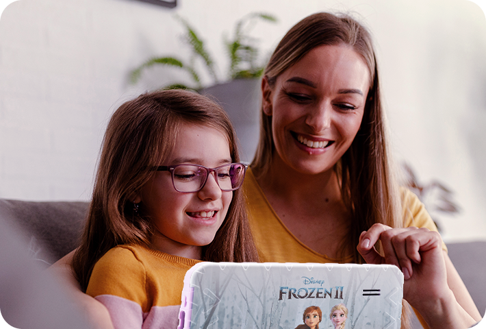 Foto de uma mãe e sua filha sentadas em um sofá, onde a criança se encontra com o Tablet Frozen em mãos. Ambas estão sorridentes.