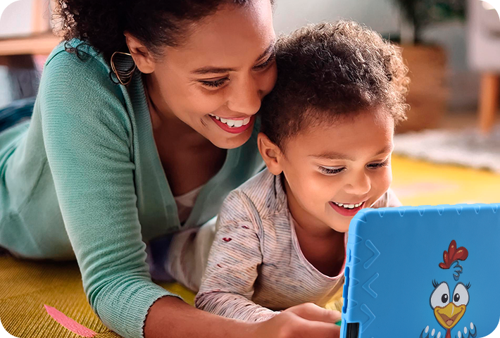 Mãe e filho deitados, em uma sala de estar, usando o Tablet Galinha Pintadinha.