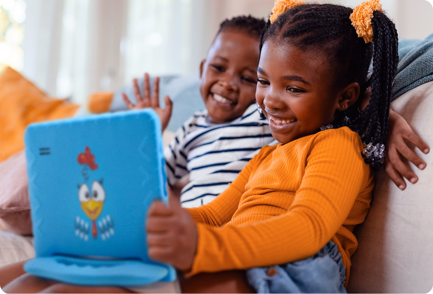 Foto de uma menina usando o Tablet Galinha Pintadinha ao lado de um garoto; ambos estão sentados em um sofá e sorrindo.